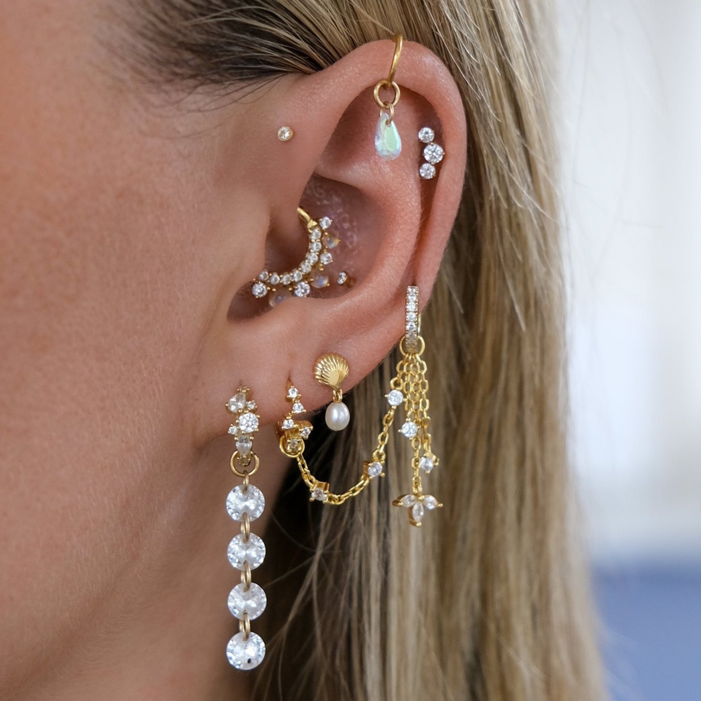 A close-up of a layered ear stack features multiple piercings with elegant gold and pearl earrings, including chains, hoops, and studs. The LANI Marquise Droplet Chain Charm by Crystal Heaven London adds a sophisticated touch.