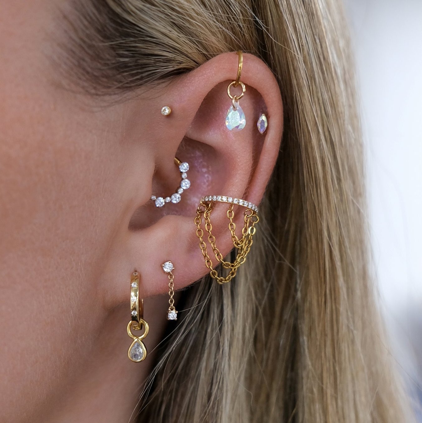 An ear adorned with gold and sterling silver hoops, studs, and chain earrings set with small gems. Among the cubic zirconia accents is the sparkling Teardrop Bezel Charm by Crystal Heaven London.