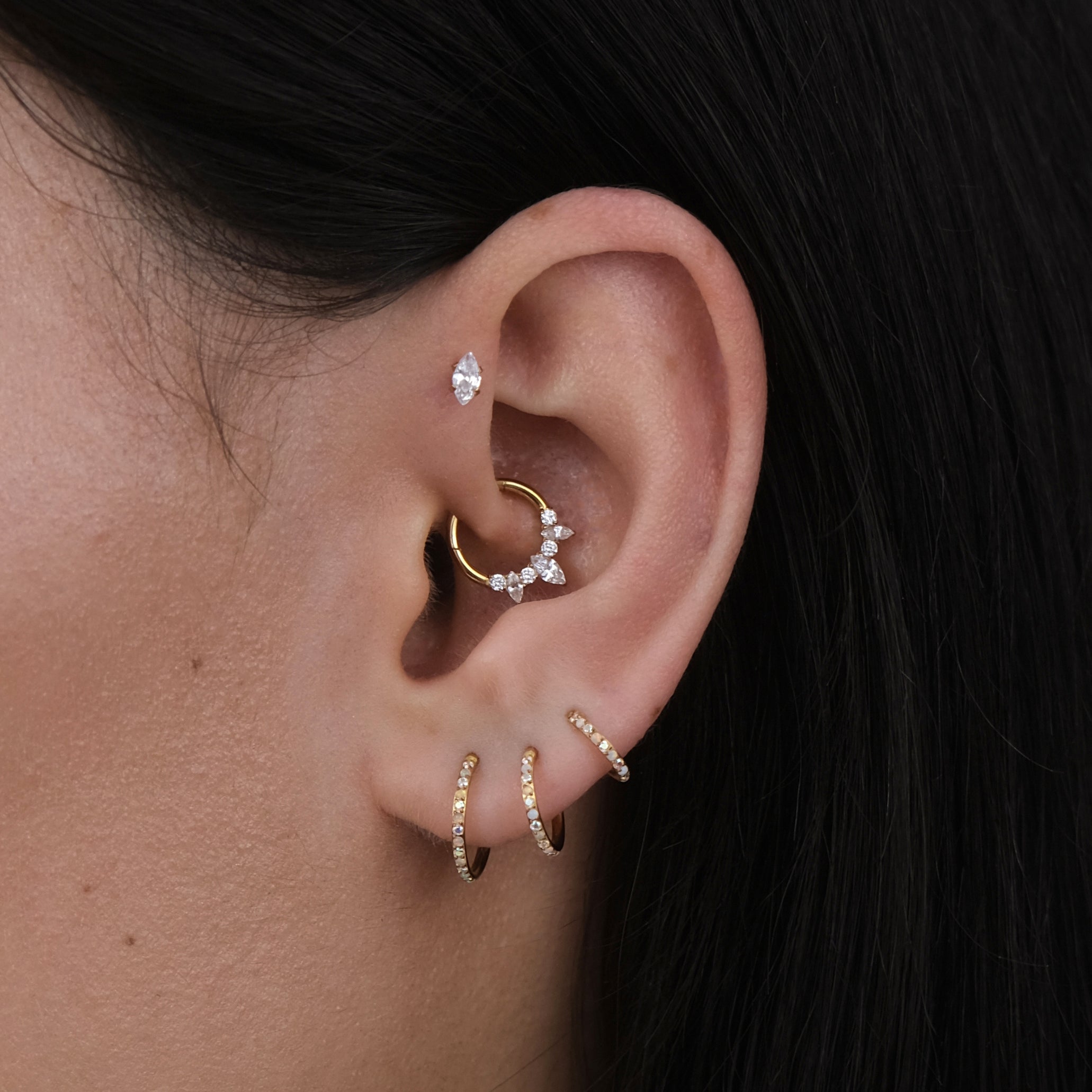 A close-up of a girls ear with multiple cartilage piercings and gold jewellery, including a crystal daith hoop.