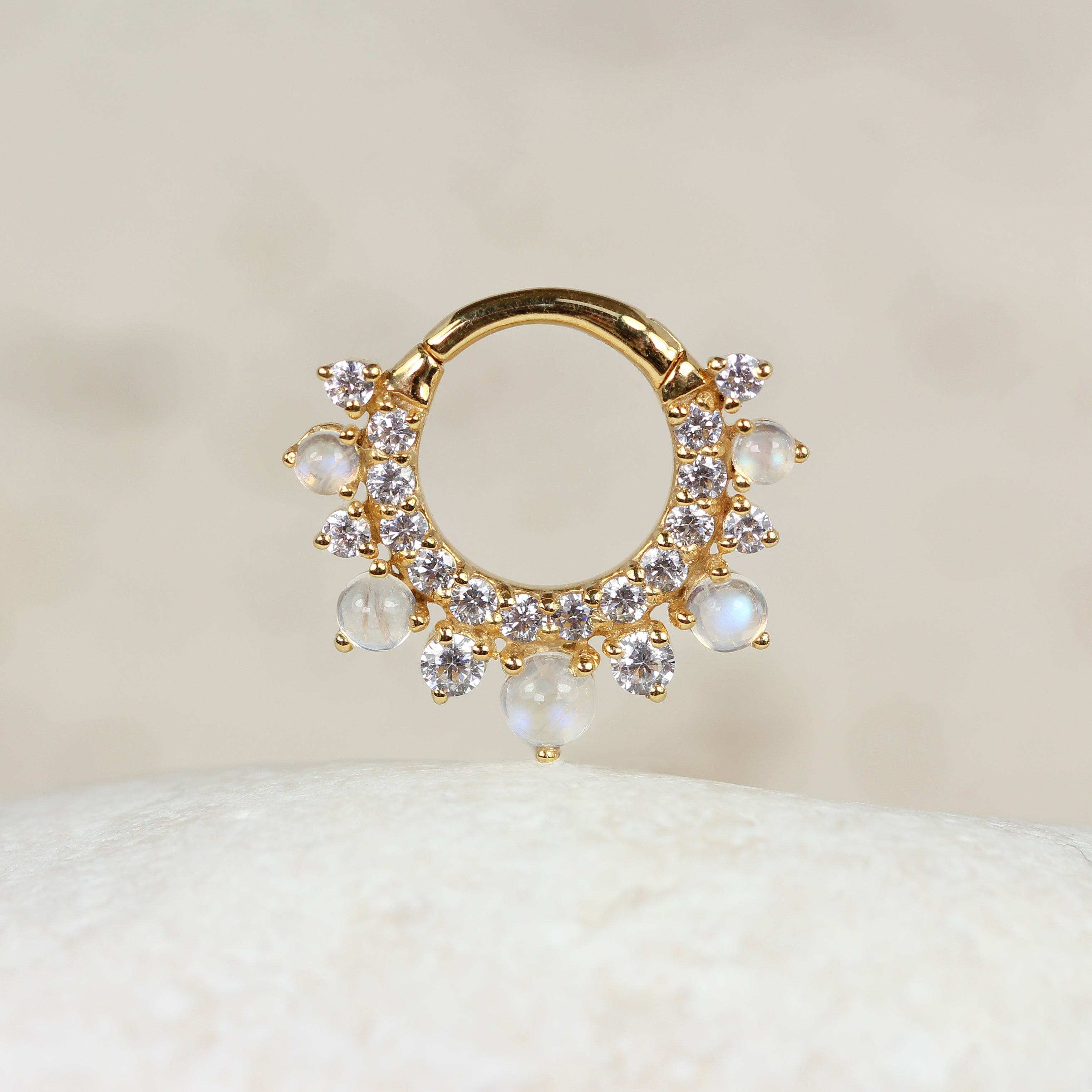 A CHLUXE CALYPSO Moonstone Daith Hoop in 14k Yellow Gold by Crystal Heaven London, embellished with delicate stones, stands vertically on a smooth white surface.